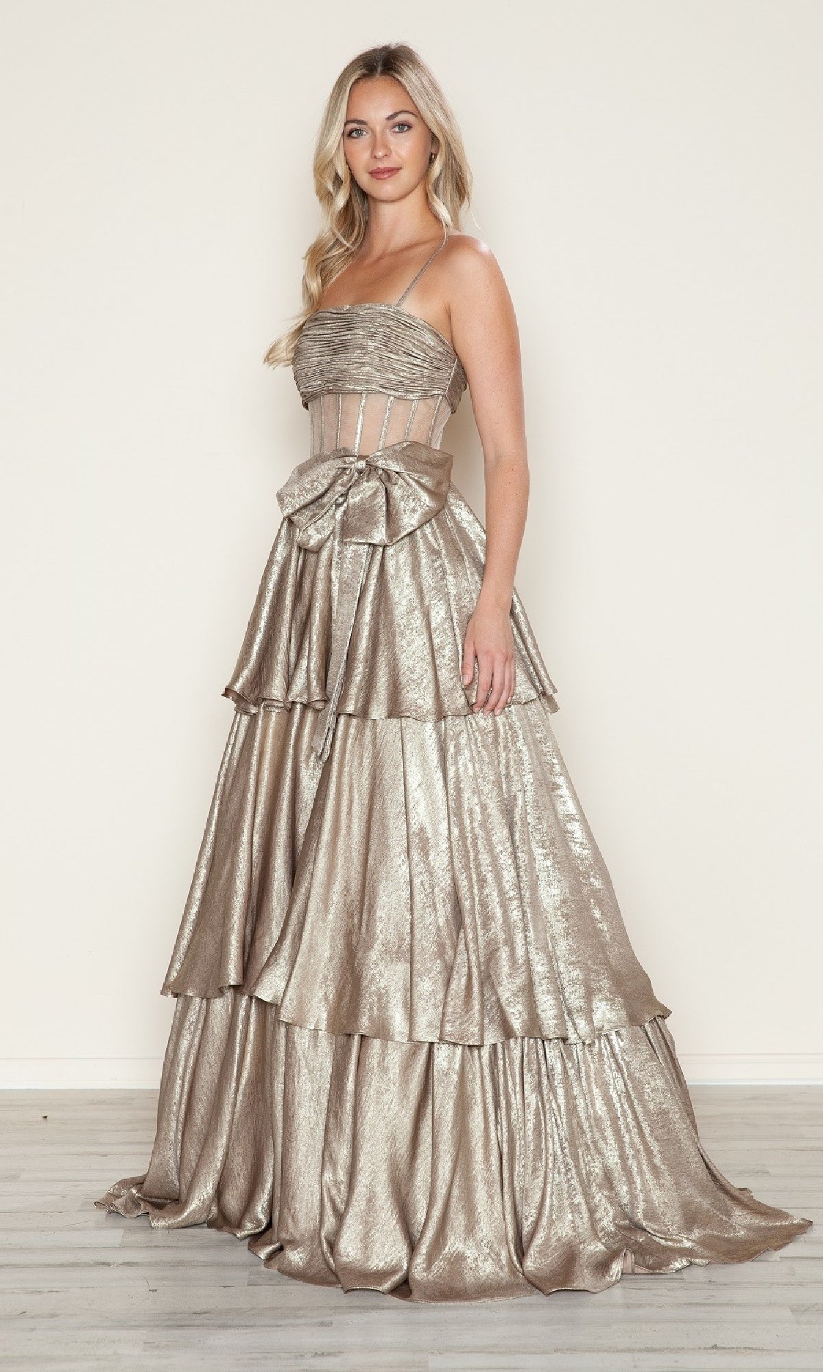 A woman models the Long Prom Dress: Poly USA 9718, featuring metallic fabric, a tiered A-line skirt, thin straps, visible boning on the fitted bodice, and a large waist bow. She stands on light wood flooring against a neutral background.